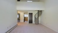 This two-floor space makes a striking first impression with its soaring double-height ceilings and open vertical volume, creating a sense of generous, airy scale. A dramatic wrought-iron spiral staircase serves as a functional centrepiece, elegantly connecting the main level to the upper loft-style floor, which overlooks the living area below via an open-railing mezzanine. Natural light filters in from above through a skylight, further enhancing the bright, open atmosphere of the space.

The main level features neutral-toned carpet throughout the living area, with a baseboard heating unit running along the perimeter wall. A dark-panelled door adds a bold contrast to the light walls and provides access to an adjacent room or storage area. Beyond the main living space, the layout opens into a kitchen, which is equipped with warm wood cabinetry, a stainless steel refrigerator, and a window that brings in additional natural light. The overall palette of soft neutrals and light tones throughout both levels contributes to a clean, understated ambiance well-suited for a variety of interior styles.