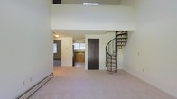 This two-floor space makes a striking first impression with its soaring double-height ceilings and open vertical volume, creating a sense of generous, airy scale. A dramatic wrought-iron spiral staircase serves as a functional centrepiece, elegantly connecting the main level to the upper loft-style floor, which overlooks the living area below via an open-railing mezzanine. Natural light filters in from above through a skylight, further enhancing the bright, open atmosphere of the space.

The main level features neutral-toned carpet throughout the living area, with a baseboard heating unit running along the perimeter wall. A dark-panelled door adds a bold contrast to the light walls and provides access to an adjacent room or storage area. Beyond the main living space, the layout opens into a kitchen, which is equipped with warm wood cabinetry, a stainless steel refrigerator, and a window that brings in additional natural light. The overall palette of soft neutrals and light tones throughout both levels contributes to a clean, understated ambiance well-suited for a variety of interior styles.