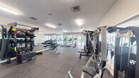 This fitness center is a comprehensive, single-floor facility designed to support a wide range of workout routines. The space is laid out with dedicated zones for cardio, strength training, and functional fitness. Durable rubber tile flooring covers the entire area, providing both safety and comfort during exercise.

The cardio section features multiple treadmills, an elliptical trainer, a stationary bike, and a rowing machine, all positioned to take advantage of natural light streaming through large glass-panel doors and windows that overlook an outdoor area. This connection to the outdoors creates an open, airy atmosphere throughout the space.

Along one wall, a broad floor-to-ceiling mirror visually expands the room while allowing users to monitor their form. Adjacent to the mirror is a well-stocked free weight area with a full rack of dumbbells, medicine balls, stability balls, and plyometric boxes, catering to varied training needs. A multi-functional cable machine and additional strength training stations round out the equipment offerings.

Overhead, bright recessed and flush-mount lighting ensures the entire space is evenly and well-lit. The clean white walls and neutral color palette contribute to a focused, professional environment. A water station is also present, adding a practical amenity for users. Overall, this fitness center provides a fully outfitted, functional space suitable for all fitness levels.