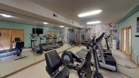 This fitness center offers a comprehensive workout environment designed to accommodate a range of exercise needs. The space is finished with soft teal-green walls and warm wood trim accents, creating a welcoming atmosphere complemented by bright overhead fluorescent lighting that ensures excellent visibility throughout.

The cardio zone is prominently featured along one side of the room, housing multiple Life Fitness treadmills, elliptical trainers, and a stationary bike. A wall-mounted flat-screen television is positioned for easy viewing during workouts. Floor-to-ceiling mirrors line one full wall, visually expanding the space and allowing users to monitor their form during exercise.

A dedicated strength training area features a cable and pulley multi-functional machine, TRX suspension straps, and a stability ball, providing options for functional and resistance-based training. A striking comic book-inspired mural wallpaper covers the accent wall in this zone, adding a bold and energetic visual element that sets a motivating tone.

The facility also includes a snack and beverage vending machine near the entrance, offering convenient refreshment access. Windows with wood-framed blinds allow for natural light while maintaining privacy. The light-colored flooring and a blue exercise mat runner contribute to a clean, practical layout. The overall design balances functionality with a lively, energetic ambiance.