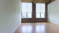 This two-floor space showcases an industrial loft aesthetic defined by a thoughtful blend of raw and refined materials. The main level is anchored by warm hardwood flooring that reflects natural light pouring in through a series of large, multi-pane windows fitted with clean white roller shades for adjustable privacy. The windows span generously from mid-wall to near the ceiling, emphasizing the impressive vertical scale of the space.

Exposed brick walls provide rich texture and character, acting as a striking architectural backdrop throughout. Overhead, the ceiling features exposed wooden beams and structural elements that reinforce the building's original industrial heritage while adding warmth to the open layout.

The combination of white-plastered walls alongside the natural brick creates a balanced contrast between contemporary and historic design elements. The open floor plan offers excellent flexibility for a variety of uses and configurations. With soaring ceiling heights, abundant natural light, original industrial details, and two full floors, this space delivers a compelling and versatile environment with strong architectural character.