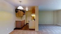 This single-floor residence offers a practical and comfortable layout designed for everyday living. The kitchen is a central feature, showcasing warm honey-toned wood cabinetry that extends to the upper walls, providing ample storage space. A decorative pendant light hangs overhead, complementing the flush-mount ceiling fixture and adding a welcoming ambiance to the space. The kitchen is equipped with a white gas range and a full-size refrigerator, with a under-cabinet range hood positioned above the cooking area. Light-toned countertops contrast nicely with the rich dark hardwood flooring that runs throughout the kitchen, giving the space a grounded, cohesive look. A peninsula-style counter provides additional workspace and a natural transition point between the kitchen and the adjoining living area. The living area features neutral wall-to-wall carpeting, creating a softer, more relaxed atmosphere in contrast to the kitchen's hard flooring. Smooth, neutral-toned walls throughout the residence contribute to a clean and airy feel. The open sightline between the kitchen and living area enhances the sense of space, making the overall layout feel connected and functional.