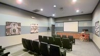 This single-level screening and presentation room is designed to accommodate small group viewings or meetings in a cinema-inspired setting. The space features rows of dark, cushioned theater-style chairs arranged in a tiered layout facing a large wall-mounted projection screen, with a ceiling-mounted projector providing a dedicated viewing setup. The walls are painted in layered bands of gray and sage green, contributing a modern, understated aesthetic. Recessed ceiling lights provide even overhead illumination throughout the room. A built-in wooden credenza or cabinet runs along the wall beneath the projection screen, offering functional storage or display space. A playful "Picture Start" film clapperboard artwork mounted on the side wall reinforces the cinematic theme of the space. The ceiling is finished in a deep charcoal gray, adding depth and contrast to the lighter walls. A doorway along the side wall provides access to the room. Overall, the space is thoughtfully arranged to create an intimate, functional environment suitable for film screenings, presentations, or private group viewings.