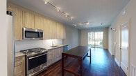 This contemporary single-floor unit showcases a thoughtfully designed open-concept layout that seamlessly connects the kitchen and living areas. The kitchen is a standout feature, outfitted with light wood grain cabinetry extending to the ceiling, stainless steel appliances including a gas range, built-in microwave, dishwasher, and refrigerator, paired with clean white countertops and a subway tile backsplash. A large island with a dark countertop provides additional workspace and dining potential. Track lighting runs across the ceiling, offering versatile illumination throughout the space. Rich, dark hardwood flooring flows continuously through the kitchen, dining, and living areas, adding warmth and visual continuity. The living area is bright and open, leading directly to a private balcony through a large glass door framed by a grid-patterned railing, allowing natural light to pour in and offering pleasant outdoor views. A generously sized bedroom features a large picture window overlooking a landscaped exterior, flooding the room with natural light. The bathroom is finished with light tile flooring, a wood-toned vanity with a framed mirror, and a glass-enclosed shower, presenting a clean and modern aesthetic throughout.