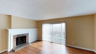 This multi-floor space showcases a welcoming living area characterized by warm hardwood flooring that extends throughout the open layout. A textured ceiling adds visual depth overhead, while a pendant light fixture provides a central focal point in the main living space. The room is anchored by a classic brick fireplace with a white-painted surround, offering a traditional design element that adds character and warmth to the area.

Natural light flows generously into the space through large sliding glass doors dressed with vertical blinds, which open onto a balcony offering views of the surrounding exterior. The neutral, warm-toned walls create a versatile backdrop that complements the rich wood tones of the flooring. A structural beam detail subtly divides the living space, adding architectural interest while maintaining an open, cohesive feel.

The overall ambiance is clean and understated, with classic finishes throughout. The combination of the fireplace, generous natural light, and balcony access gives the living area a comfortable, functional character. Spanning three floors, the property offers a substantial amount of living space across multiple levels.