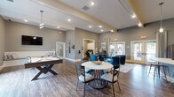 Located at 100 Legends Dr in Canton, GA, this single-floor community clubhouse offers a well-appointed gathering space designed for both relaxation and productivity. The main lounge area is expansive and open, featuring soaring ceilings, recessed lighting, and rich hardwood-style flooring throughout. Neutral gray walls are complemented by bold teal upholstered seating, a round central table, and accent rugs that define distinct activity zones within the open-plan layout.

A dedicated billiards room provides a full-size pool table, a wall-mounted flat-screen TV, and a built-in bench with cushioned seating — an ideal space for casual recreation. Adjacent to the main lounge, a business center offers dual computer workstations with white built-in cabinetry, a printer, and framed artwork, making it a functional workspace for residents.

The kitchenette is equipped with white shaker-style cabinetry, quartz countertops, a stainless steel dishwasher, a full-size refrigerator, and a Starbucks coffee machine — providing a premium café-style amenity. Glass double doors at the rear of the space open directly to an outdoor pool and patio area, flooding the interior with natural light and offering seamless indoor-outdoor connectivity.