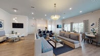 This thoughtfully designed single-floor space offers a bright and welcoming environment highlighted by a cohesive navy, cream, and warm neutral color palette. The expansive main lounge area serves as the heart of the space, featuring light hardwood-style flooring, crisp white walls with crown molding, and a tray ceiling with a soft blue accent. A statement glass chandelier anchors the central seating arrangement, which includes plush sofas and armchairs upholstered in patterned blue fabric, centered around a wood coffee table. Bold abstract artwork adorns the walls, adding a curated, contemporary touch. Adjacent to the lounge, a dedicated dining area is bathed in natural light streaming through large windows dressed with bold geometric-patterned drapes and shuttered blinds, offering views of lush outdoor greenery. Round dining tables paired with upholstered chairs create an inviting space for gathering. A kitchenette tucked along one wall features white cabinetry, a stainless steel sink and appliances, and striking starburst wall sconces that illuminate a textured white tile backsplash. Additional seating nooks throughout the space provide flexible areas for smaller gatherings or quiet use. The open, airy layout, abundant natural light, and refined furnishings combine to create a polished yet comfortable communal environment.