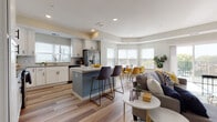 This well-appointed single-floor residence offers a thoughtfully designed open-concept layout that seamlessly connects the kitchen, dining, and living areas. The kitchen is a standout feature, showcasing crisp white cabinetry, granite countertops, a classic subway tile backsplash, stainless steel appliances, and a blue-accented island with bar seating — ideal for both meal preparation and casual dining. Light wood-style flooring flows throughout the main living areas, enhancing the bright and airy atmosphere created by abundant natural light streaming through multiple windows. The dining nook is positioned in a sun-filled corner with large windows offering outdoor views, creating a welcoming space for entertaining. The living room is generously proportioned, featuring vaulted ceilings, soft neutral tones, and a sliding glass door that opens to a private balcony. The bedroom is a serene retreat, carpeted for comfort, with large windows dressed in sheer curtains that invite natural light. A spacious walk-in closet with wire shelving provides ample storage. The bathroom features wood-look tile flooring, a granite-topped white vanity with matte black hardware, and a full tub-shower combination. A dedicated in-unit laundry room with a full-size washer and dryer adds everyday convenience.