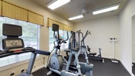 This fitness center is a dedicated single-floor space designed to support a well-rounded exercise routine. The room is bright and welcoming, benefiting from multiple windows dressed with yellow valances and horizontal blinds that allow ample natural light to filter in, offering pleasant views of the surrounding greenery outside. Overhead, recessed fluorescent lighting and a ceiling fan ensure the space remains well-lit and comfortable during workouts. The flooring is finished in dark, durable material suited for high-traffic gym use. The equipment selection is comprehensive, including an elliptical trainer, a stationary cycle, and a multi-function cable and weight machine, along with additional strength training stations positioned throughout the room. The walls are painted in a clean, neutral tone, contributing to an open and airy atmosphere. The thoughtful arrangement of equipment allows for ease of movement between machines, making the space efficient and user-friendly. Overall, this fitness center provides a practical and inviting environment for cardiovascular conditioning and strength training alike.