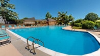 Located at 211 Lingstorm Lane in North Chesterfield, Virginia, this outdoor recreational facility centers around a generously sized freeform swimming pool with clearly marked depth indicators. The pool's vivid blue water is complemented by a broad, open concrete sundeck that wraps around the perimeter, offering ample space for relaxation and movement.

The deck is well-appointed with numerous teal lounge chairs and umbrella-shaded tables, providing both sunny and shaded options for guests. A raised terrace area, framed by a distinctive natural stone retaining wall, overlooks the pool and features additional seating beneath bright green umbrellas, creating a casual gathering space with elevated views of the water.

The entire pool area is enclosed by a sturdy wrought iron fence with a gated entry, ensuring safety and privacy. Prominent signage at the entrance outlines pool rules and regulations. A utility shed and support building with a pitched roof are situated adjacent to the pool, offering functional amenities for the facility.

Mature trees surround the perimeter, providing natural shade and a lush, park-like ambiance. Manicured landscaping with shrubs, mulched beds, and green open lawn areas further enhance the outdoor setting, making this a well-maintained and inviting community recreational destination.