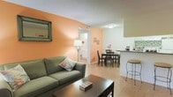 This single-story space is characterized by its warm, inviting ambiance, highlighted by bold peach-toned accent walls that add a vibrant yet welcoming energy throughout. The living area serves as the central focus, featuring light hardwood-style flooring that extends seamlessly into the adjoining spaces, creating visual continuity. A comfortable seating area anchors the room, complemented by a dark wood coffee table and a floor lamp that contributes to the overall warmth of the space. The open layout allows for a natural flow between the living room and the dining area, where a dark wood dining set provides a defined space for meals. Adjacent to the dining area, a kitchen counter with bar-style seating offers an additional casual dining option, with the kitchen positioned conveniently nearby. A textured ceiling adds architectural detail overhead. A doorway visible from the living area leads to a bathroom, suggesting a practical and well-connected floor plan. Natural light filters into the space, enhancing the bright, airy quality of the interior. Overall, the space balances functionality and comfort through its cohesive design elements.