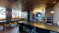 This striking single-floor space masterfully blends rustic and contemporary design elements to create a warm yet refined atmosphere. Throughout the interior, rich wood-paneled walls and ceilings pair seamlessly with stacked stone accents, while wide-plank hardwood flooring ties the various areas together cohesively.

The centerpiece of the space is an expansive dining and lounge area anchored by a large, dark wood communal table surrounded by leather and woven chairs. Dramatic orb-shaped chandelier pendants hang overhead, casting ambient light across the room. A built-in cushioned bench with under-seat LED accent lighting runs along the floor-to-ceiling windows, offering sweeping panoramic views of the surrounding cityscape and treetops.

The fully equipped kitchen features dark granite countertops, warm wood cabinetry, stainless steel appliances including double wall ovens, and globe pendant lighting suspended from a wood-planked ceiling. A generously sized island with bar seating provides additional gathering space and a wet bar area with a round mirror and stone backdrop adds a refined finishing touch.

A comfortable lounge nook with velvet accent chairs and open shelving opens directly to an expansive outdoor terrace through large black-framed glass doors, flooding the interior with natural light. A separate seating area with leather furnishings and a central fireplace completes this versatile and meticulously designed space.