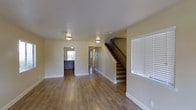 This two-story home offers a clean, well-maintained interior designed for comfortable everyday living. The main level features warm hardwood-style flooring that runs throughout, lending a cohesive and inviting feel to the open living space. Soft yellow-toned walls reflect light evenly, complemented by flush-mount ceiling light fixtures that provide consistent illumination throughout the area. A central staircase with a dark wood railing serves as a functional focal point, connecting the main floor to the upper level with an efficient and straightforward design. The layout flows naturally from the living area through to additional rooms toward the rear of the home, with doorways leading to what appears to be a kitchen or utility area, as well as a hallway providing access to further spaces. White window blinds allow for adjustable natural light, contributing to the bright and airy atmosphere of the main floor. Clean white baseboards and neutral wall tones throughout give the home a fresh, move-in-ready appearance. The overall design is practical and uncluttered, making it an adaptable space suited to a variety of living arrangements across both floors.