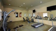 This fitness room offers a practical and well-organized space designed to support a variety of workout routines. The room features carpeted flooring throughout, providing a comfortable surface underfoot, and is illuminated by recessed ceiling lighting that ensures a bright, evenly lit environment. Neutral-toned walls contribute to a clean, focused atmosphere suited for exercise.

The space is equipped with a selection of cardio and strength training machines, including a treadmill, an elliptical cross-trainer, a stationary recumbent bike, and an upright cycle. A weight bench is positioned in the foreground, offering additional options for strength training. A wall-mounted flat-screen television is installed above the mirror, allowing residents to stay entertained during workouts.

A large mirror spans one wall, visually expanding the space and allowing users to monitor their form during exercise. Framed artwork decorates the walls, adding a touch of warmth to the otherwise functional setting. Double doors with glass panels lead to an adjacent area, allowing natural light to filter into the room and providing a sense of connection to the surrounding environment.