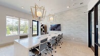 Located in Pensacola, FL, this thoughtfully designed office space offers a refined, contemporary environment suited for professional use. The main conference area serves as the centerpiece, anchored by a long white marble-topped table surrounded by ergonomic mesh chairs. Two striking geometric pendant light fixtures with globe bulbs and wood-toned frames hang overhead, adding a modern yet warm aesthetic to the space. A large wall-mounted display screen is set against a subtly textured accent wall, providing an ideal setup for presentations and meetings. Neutral-toned carpet tile flooring runs throughout, complementing the crisp white walls and bright, airy atmosphere. A bank of expansive windows along one wall floods the space with natural light while offering views of the surrounding greenery and palm trees. Black-framed glass doors lead to additional private office spaces, each featuring botanical-motif frosted glass panels and individual workstations. A built-in white cabinetry unit with a teal accent shelf provides functional storage and display space, while wall sconces add a warm, finishing touch. The overall design balances clean modern lines with inviting accents, creating a professional yet comfortable workspace.