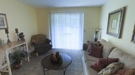This single-floor residence offers a comfortable and functional layout designed for relaxed everyday living. The living room serves as the heart of the home, featuring soft yellow walls that lend a warm and cheerful ambiance to the space. A large sliding glass door dressed with vertical blinds acts as a central focal point, allowing an abundance of natural light to pour in while providing access to the outdoors. The room is finished with plush wall-to-wall carpeting underfoot, adding a sense of warmth and comfort. The space is well-proportioned, comfortably accommodating a sofa adorned with decorative pillows, a cozy armchair, and a round coffee table that anchors the seating arrangement. A decorative vase with floral accents adds a touch of elegance to the room. The textured ceiling completes the classic residential aesthetic of the interior. Overall, the space presents a clean, inviting atmosphere that is both practical and homey, making it well-suited for comfortable day-to-day living.
