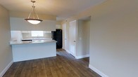 This single-floor space offers a clean, functional layout designed to maximize everyday living. The open-concept design seamlessly connects the kitchen and living areas, creating a cohesive and airy feel throughout. The kitchen is equipped with a peninsula-style counter that doubles as a workspace and casual dining surface, complemented by white cabinetry, a built-in sink with a window above it, and a black refrigerator. A stylish pendant light hangs above, adding a warm focal point to the space. Rich, dark hardwood-style flooring runs throughout, providing a grounded contrast to the soft, neutral beige walls and crisp white baseboards. The layout includes multiple interior doors leading to additional rooms or storage areas, offering a well-organized flow from one space to the next. Flush HVAC vents integrated into the floor and walls indicate a central climate control system. The overall ambiance is bright, clean, and move-in ready, with a practical design that suits a variety of living arrangements. The consistent finishes and open sightlines throughout give the space a unified, well-maintained character.