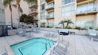 Located at 5745 Case Ave in North Hollywood, this outdoor amenity area is part of a modern mid-rise building distinguished by its contemporary architectural design. The facade features large floor-to-ceiling windows, glass-railed private balconies, and a warm neutral stucco exterior accented by tropical palm plantings throughout the courtyard.

The centerpiece of the space is a generously sized rectangular swimming pool with clear, light-catching water, surrounded by a spacious tiled sun deck furnished with multiple lounge chairs and patio umbrellas for shade. Adjacent to the pool sits an octagonal spa, complemented by additional seating areas including cushioned outdoor sofas and side tables, creating a comfortable social environment for relaxation.

Raised planters with lush tropical vegetation frame the perimeter of the courtyard, adding a resort-like atmosphere. An elevated sun terrace extends beyond the pool area, offering panoramic views of the North Hollywood skyline, surrounding treetops, and distant mountain ranges under the characteristic Southern California blue sky. The open-air setting benefits from abundant natural light throughout the day, contributing to the inviting, leisure-oriented ambiance of the space.