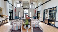 This single-floor community space makes an immediate impression with its dramatic, soaring ceilings and generous use of natural light. The grand lobby-style entrance area features towering columns, bold dark trim accents, and a series of tall arched windows — including a prominent arched transom above the main entry doors — that flood the space with daylight. Patterned wingback chairs and striped accent pillows are arranged around a central pedestal table adorned with a vibrant floral arrangement, creating a welcoming and refined seating area.

Beyond the entrance, the space transitions into an expansive lounge with warm hardwood-style flooring and a striking floor-to-ceiling stone fireplace as the focal point. A reclaimed wood surround frames a mounted flat-screen television above the firebox, and large windows on either side bring in abundant natural light while offering views of the surrounding greenery. Comfortable leather seating and bistro-height tables provide versatile areas for relaxing or socializing.

Adjacent to the lounge, a well-appointed kitchenette offers modern grey cabinetry, open shelving with decorative plants, a stainless steel refrigerator, and a coffee and beverage station — ideal for hosting community events or casual gatherings. The overall ambiance blends classic architectural grandeur with contemporary comfort throughout.