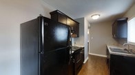 This single-floor space showcases a kitchen designed with a cohesive, contemporary aesthetic. Dark espresso cabinetry lines the kitchen walls, providing ample storage and a bold visual contrast against the neutral grey walls. The kitchen is equipped with matching black appliances, including a full-sized refrigerator, an electric range with a built-in overhead microwave, and additional counter space visible toward the back. Rich, dark hardwood-style flooring runs throughout, adding warmth and continuity to the space. The textured ceiling and flush-mount ceiling light provide functional illumination. A hallway extends beyond the kitchen, hinting at additional rooms within the layout. White baseboards frame the flooring neatly, offering a clean finishing detail throughout. The overall ambiance is modern and understated, with the dark tones of the cabinetry and appliances creating a striking, unified look that is both practical and visually appealing.