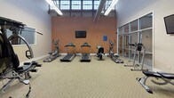 This fitness center is a well-appointed, single-floor facility divided into several distinct zones, each designed to support a comprehensive workout experience. The standout feature is the expansive indoor basketball court, finished with a two-tone modular sport-tile floor in green and grey, complete with painted court markings and regulation backboards at each end. A custom logo at center court adds a distinctive touch. Soaring ceilings with exposed dark wood timber trusses rise overhead, flanked by rows of large industrial-style clerestory windows that flood the court with abundant natural light.

Adjacent to the court, a dedicated cardio area houses treadmills, elliptical trainers, and a stationary bike, all arranged beneath similarly high ceilings with clerestory windows. A wall-mounted television provides entertainment, while glass partitions offer visual connectivity between zones. A separate strength training room features a multi-station cable machine, a full dumbbell rack, a medicine ball, and a barbell, set on durable patterned flooring. An additional open carpeted room offers flexible space for stretching or group activities. Neutral-toned walls, clean white baseboards, and consistent flooring throughout unify the space with a polished, functional aesthetic.