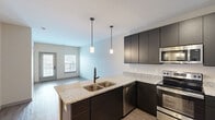 Located at 316 Main St in Hamilton, Ohio, this one-bedroom, one-bathroom unit offers a thoughtfully designed single-floor layout with modern finishes throughout. The open-concept kitchen and living area serves as the heart of the space, where the kitchen stands out with bold dark cabinetry reaching to the ceiling, light granite countertops, a peninsula with a double stainless steel sink, and pendant lighting overhead. A full suite of stainless steel appliances — including a refrigerator, electric range, built-in microwave, and dishwasher — completes the kitchen. Light wood-tone flooring runs throughout the main living area, lending a clean and contemporary feel. The living room receives natural light through a window overlooking an exterior balcony railing, creating a bright and welcoming atmosphere. The bedroom features plush carpeting, neutral walls, and a sliding door closet with built-in shelving, providing functional storage. The bathroom mirrors the kitchen's design aesthetic with dark cabinetry, granite countertops, a large mirror, and stylish light fixtures above the vanity, complemented by wood-look tile flooring and a separate shower area.