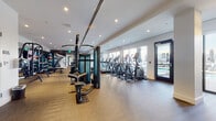This contemporary fitness center is designed to offer a comprehensive workout experience within a thoughtfully laid-out, single-floor space. The facility is flooded with natural light through large floor-to-ceiling windows that line the perimeter, offering views of an adjacent outdoor pool area accessible via glass-paneled doors.

The main workout floor features herringbone-patterned carpet throughout and is divided into distinct functional zones. A dedicated cardio section lines the window wall, housing a row of treadmills, elliptical trainers, and stationary cycling bikes, along with a large display screen to keep users engaged during workouts. Matrix-branded cable machines and functional training stations anchor the central zone, providing versatile strength training options.

A separate strength and free weights area includes a heavy-duty squat rack loaded with Olympic plates, a full dumbbell rack, and additional benching equipment, all reflected in floor-to-ceiling mirrors that amplify the sense of space. Built-in open shelving with wood accents provides organized storage for accessories and amenities.

Recessed lighting throughout ensures consistent illumination across all workout zones, while the clean white walls and neutral tones create a bright, professional atmosphere. The space also features a posted studio rules board, indicating its managed, community-use nature. Direct outdoor access reinforces a seamless indoor-outdoor lifestyle experience.