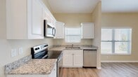 This single-floor unit offers a well-designed, open layout that maximizes functionality and comfort. The kitchen serves as the centerpiece of the space, featuring crisp white shaker-style cabinetry extending to the upper walls, speckled granite countertops, and a full suite of stainless steel appliances including a refrigerator, electric range with over-the-range microwave, and dishwasher. A double-basin sink is positioned beneath a window fitted with horizontal blinds, allowing natural light to brighten the workspace. The kitchen flows seamlessly into an open living area distinguished by soaring vaulted ceilings that lend a sense of spaciousness well beyond the unit's compact footprint. A ceiling fan is centrally mounted, providing both air circulation and light. Light wood-tone luxury vinyl plank flooring runs throughout the entire space, contributing to a clean, cohesive aesthetic. Warm beige walls complement the neutral palette, while multiple windows dressed with blinds invite ample natural light from multiple directions. The open living area provides flexible use of space, with clear sightlines connecting the kitchen and main living zones. The overall ambiance is bright, modern, and move-in ready.