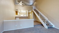 The Clipper is a thoughtfully designed two-story residence offering one bedroom and one bathroom across a generous floor plan. The main level features light hardwood-style flooring throughout the entry and kitchen areas, transitioning to plush carpet in the living room. The kitchen is equipped with white cabinetry accented by warm wood trim, a suite of white appliances, ample counter space, and a peninsula that opens to the living area — ideal for everyday functionality. The living room is a standout space, boasting soaring ceilings, a ceiling fan, a tile-surround fireplace, and a sliding glass door that leads to a private balcony with outdoor views. A staircase with white railings rises to the upper level, where the carpeted bedroom offers a sliding door to its own balcony, a large window for natural light, and a spacious closet with built-in shelving. The upper hallway overlooks the main level below, enhancing the open, airy feel of the home. The full bathroom includes a single-sink vanity with a speckled countertop, white cabinetry, and a full tub-shower combination. Throughout, warm neutral tones create a cohesive and comfortable ambiance.