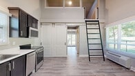 Located at 2027 W Powell Blvd in Gresham, Oregon, this thoughtfully designed one-bedroom, one-bathroom unit makes excellent use of its compact footprint. The open-concept living and kitchen area immediately impresses with soaring vaulted ceilings and a loft space accessible via a sleek black ladder, adding vertical dimension and architectural interest. The kitchen is well-appointed with dark cabinetry, white countertops, and a full suite of stainless steel appliances including a range, dishwasher, refrigerator, and microwave. A stylish sliding barn door connects the main living area to the bedroom corridor, adding a modern farmhouse touch. Light gray luxury vinyl plank flooring runs throughout the entire unit, providing a clean, cohesive look. The bedroom is a comfortable, private retreat featuring a closet, built-in shelving nooks, and a window that brings in natural light. The bathroom offers a tile-surround walk-in shower, a white vanity with mirror, and a window for ventilation and natural light. Multiple windows throughout the unit frame views of surrounding greenery, keeping the interior bright and connected to the outdoors. The overall ambiance is clean, modern, and move-in ready.