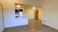 This single-floor space offers a clean, modern design with an open-concept layout that seamlessly connects the kitchen and living areas. The kitchen is equipped with stainless steel appliances, including a full-size refrigerator, and features dark cabinetry that contrasts neatly against light countertops. A peninsula with a built-in sink provides additional workspace and acts as a natural divider between the kitchen and the open living area, making it ideal for casual dining or entertaining. Warm hardwood-style flooring runs throughout the main living space, lending a cohesive and inviting feel to the interior. The walls are finished in a soft, neutral tone that enhances the brightness of the space, complemented by flush-mount ceiling lighting that provides even illumination. A carpeted area is visible at the edge of the living space, suggesting a defined lounge or relaxation zone. A doorway leads to additional rooms, offering a sense of privacy and separation. The layout is functional and efficient, making excellent use of the available space while maintaining an airy, open atmosphere throughout.