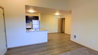 This single-floor space offers a clean, modern design with an open-concept layout that seamlessly connects the kitchen and living areas. The kitchen is equipped with stainless steel appliances, including a full-size refrigerator, and features dark cabinetry that contrasts neatly against light countertops. A peninsula with a built-in sink provides additional workspace and acts as a natural divider between the kitchen and the open living area, making it ideal for casual dining or entertaining. Warm hardwood-style flooring runs throughout the main living space, lending a cohesive and inviting feel to the interior. The walls are finished in a soft, neutral tone that enhances the brightness of the space, complemented by flush-mount ceiling lighting that provides even illumination. A carpeted area is visible at the edge of the living space, suggesting a defined lounge or relaxation zone. A doorway leads to additional rooms, offering a sense of privacy and separation. The layout is functional and efficient, making excellent use of the available space while maintaining an airy, open atmosphere throughout.
