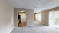 This single-floor residence is designed with a practical, comfortable layout that makes efficient use of its space. The open living area is finished with plush wall-to-wall carpeting and warm beige walls, creating an inviting atmosphere. A ceiling fan with a light fixture provides both comfort and illumination, while a large sliding glass door fitted with vertical blinds fills the room with natural light and offers views of the surrounding greenery. A secondary window further brightens the space.

The kitchen is separated from the living area by a partial wall and features warm wood-toned cabinetry, dark granite-style countertops, and hardwood-style flooring. Black appliances, including a refrigerator and dishwasher, complement the cabinetry for a cohesive look. A textured ceiling throughout the unit adds a classic residential character.

The bedrooms are generously proportioned, carpeted, and bathed in neutral tones, with large windows allowing ample natural light. The hallway connects the sleeping areas to the bathroom, which features a wood-toned vanity with a granite-style countertop and a sink. Closet doors line the hallway, offering convenient storage. The overall ambiance of the space is clean, functional, and move-in ready.