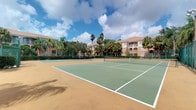 This property features a well-maintained outdoor tennis court that serves as a standout recreational amenity within a tropical residential community. The court is finished in a classic green hard-court surface with clearly marked white lines and a regulation net, providing a functional and visually appealing space for play. Surrounding the court is a perimeter fence that defines the boundaries of the playing area while maintaining an open, airy feel.

The setting is distinctly tropical, with tall palm trees and lush mature vegetation framing the court and providing a verdant backdrop. Multi-story residential buildings with warm terracotta-toned facades and tiled rooflines are visible beyond the tree line, integrating the recreational space naturally into the broader community environment. The open-sky exposure allows for abundant natural light throughout the day, enhancing both visibility and the overall outdoor experience.

The combination of the well-kept playing surface, rich landscaping, and the backdrop of the surrounding architecture creates an inviting and resort-like atmosphere. This tennis court adds significant lifestyle value to the community, offering residents a dedicated space for recreation and outdoor activity within a vibrant, Florida-inspired setting.