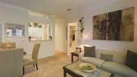 This single-floor residence offers a thoughtfully designed open-concept layout that seamlessly connects the main living spaces. The living room serves as the heart of the home, featuring plush wall-to-wall carpeting in a warm neutral tone, creating a comfortable and inviting atmosphere. A contemporary sofa and coordinating accent chairs anchor the space, while a glass-top coffee table adds a modern touch. Decorative wall art and carefully placed greenery contribute to the room's refined aesthetic.

A pass-through window connects the living area to the kitchen, promoting an open feel while maintaining defined zones. The kitchen showcases white cabinetry, a light-toned countertop, and white appliances, contributing to a clean and functional workspace. Crown molding details at the pass-through opening add an elegant architectural element.

An arched doorway leads to additional rooms beyond the living area, hinting at further private spaces within the home. Throughout, the consistent warm white wall color, neutral carpeting, and soft lighting work together to create a bright, cohesive, and welcoming environment.