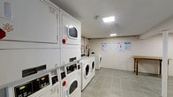 The shared laundry facility at Grandview Gardens is a well-organized, single-floor space designed to meet residents' everyday laundry needs. The room features a lineup of white stacked washer-dryer units along one wall, complemented by a row of front-loading machines that together offer ample capacity for multiple users. The machines are equipped with clearly labeled digital control panels, making operation straightforward. The floor is finished with light grey tiles that lend a clean, practical aesthetic to the space. Overhead, recessed and panel lighting provides bright, even illumination throughout the room. The walls are painted in a neutral white tone, contributing to the overall bright and tidy atmosphere. Instructional and informational notices are posted on the far wall, offering guidance to users on machine operation and laundry best practices. The layout allows for comfortable movement between machines, ensuring ease of use. Utility connections and piping are visible along the back wall, reinforcing the room's functional character. Overall, the laundry facility is a clean, efficiently arranged communal amenity.