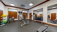 This fitness center offers a spacious, single-room layout designed to accommodate a comprehensive range of workout activities. The interior is finished with durable speckled rubber flooring throughout, providing both safety and comfort during exercise. Elegant dark wood crown molding and warm wood-trimmed windows and doors lend a refined aesthetic that sets this facility apart from a standard gym environment.

Natural light filters in through multiple windows fitted with wood-slatted blinds, while skylights and recessed ceiling panels further brighten the space. Ceiling fans promote airflow and comfort during workouts. Large wall-mounted mirrors span multiple walls, enhancing the sense of openness and allowing users to monitor their form.

The equipment selection is well-rounded, including a multi-function cable and weight machine, multiple adjustable benches, a full dumbbell rack, kettlebells, medicine balls, and additional strength training stations. A wall-mounted flat-screen television provides entertainment during workouts. Elegant French doors with glass panels serve as the main entry point, complemented by a standard door for secondary access. Signage at the entrance clearly identifies this as the Fitness Center and outlines facility rules. The overall ambiance is clean, polished, and welcoming, combining practical functionality with an upscale interior finish.