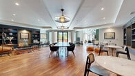 This expansive single-floor communal space offers a thoughtfully designed interior that blends mid-century modern aesthetics with warm, eclectic character. The layout flows seamlessly across several distinct functional zones, each with its own unique ambiance and purpose.

The main lounge area features warm hardwood flooring beneath a coffered ceiling with a striking pendant light fixture. Multiple seating arrangements — from modish accent chairs to intimate groupings — create flexible social spaces. A bold green barn door serves as an architectural focal point, leading into a well-appointed library nook lined with floor-to-ceiling dark navy built-in bookshelves, warm wood accents, and curated seating for quiet reading or focused work.

Transitioning into the dining and café area, the space opens dramatically with soaring ceilings and expansive floor-to-ceiling glazed walls that flood the interior with natural light and offer views of outdoor greenery. Geometric patterned tile flooring anchors communal dining tables beneath rows of industrial-style dome pendant lights. Exposed brick, a wooden ceiling installation, and a mounted flat-screen TV add character to this lively gathering zone.

Throughout, the design balances polished concrete floors, rich textiles, and curated artwork to create a vibrant yet inviting communal environment.