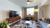 This single-floor unit showcases a cohesive mid-century modern aesthetic throughout its thoughtfully designed spaces. The living area serves as the heart of the home, featuring dark hardwood-style flooring, a ceiling fan, and large windows dressed in neutral drape panels that invite generous natural light. A dedicated dining nook with a bistro-style table sits beside the window, creating a bright casual dining space alongside a comfortable seating area.

The kitchen is well-equipped with white shaker cabinetry, light countertops, and a full suite of stainless steel appliances, including a refrigerator, range, microwave, and dishwasher. Pendant lighting and recessed fixtures provide both task and ambient illumination, while the layout connects fluidly to the living space.

Multiple bedrooms offer carpeted floors, natural light through windows with blinds and curtain panels, and neutral wall tones that provide a calm, restful atmosphere. Each room comfortably accommodates a full-sized bed and supporting furniture.

The bathrooms maintain a clean, contemporary feel, with one featuring a tub-shower combination and another offering a white vanity with a large mirror, wood-look flooring, and crisp white finishes throughout.