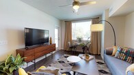 This single-floor unit showcases a cohesive mid-century modern aesthetic throughout its thoughtfully designed spaces. The living area serves as the heart of the home, featuring dark hardwood-style flooring, a ceiling fan, and large windows dressed in neutral drape panels that invite generous natural light. A dedicated dining nook with a bistro-style table sits beside the window, creating a bright casual dining space alongside a comfortable seating area.

The kitchen is well-equipped with white shaker cabinetry, light countertops, and a full suite of stainless steel appliances, including a refrigerator, range, microwave, and dishwasher. Pendant lighting and recessed fixtures provide both task and ambient illumination, while the layout connects fluidly to the living space.

Multiple bedrooms offer carpeted floors, natural light through windows with blinds and curtain panels, and neutral wall tones that provide a calm, restful atmosphere. Each room comfortably accommodates a full-sized bed and supporting furniture.

The bathrooms maintain a clean, contemporary feel, with one featuring a tub-shower combination and another offering a white vanity with a large mirror, wood-look flooring, and crisp white finishes throughout.
