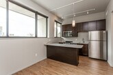 This well-appointed single-floor space showcases a contemporary design with a cohesive blend of modern finishes and functional layout. The kitchen serves as a central highlight, featuring rich dark wood cabinetry that extends to the ceiling, a striking marble-topped peninsula that doubles as a breakfast bar, and a full suite of stainless steel appliances including a gas range, built-in microwave, and refrigerator. Pendant lighting hangs above the peninsula, complementing the track lighting system that runs throughout the space. Large, black-framed windows in the kitchen frame urban views and flood the area with natural light. Warm-toned hardwood flooring flows seamlessly across the entire unit, creating a sense of continuity and warmth. The open living area adjacent to the kitchen offers generous wall space and soaring ceilings, providing a versatile canvas for a variety of furniture arrangements. A dedicated bedroom provides a quiet retreat, featuring the same hardwood flooring and neutral wall tones. The bathroom is finished with dark wood cabinetry, a marble-topped vanity, and clean, minimalist fixtures, maintaining the refined aesthetic found throughout the rest of the space.