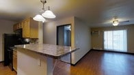 This single-floor space offers an open-concept layout that connects the kitchen and living areas in a practical, cohesive design. The kitchen features a prominent peninsula breakfast bar with a dark granite-style countertop, providing both prep space and casual seating. Oak cabinetry with a warm finish lines the upper kitchen walls, complemented by a tile backsplash that adds texture. A dual pendant light fixture with classic white shades hangs above the bar, providing focused task lighting.

Rich, warm-toned hardwood flooring extends throughout the main living area, grounding the space and offering visual continuity. The living area is equipped with a wall-mounted air conditioning unit and a baseboard heater, ensuring year-round climate control. A door with a glass panel allows natural light to filter into the space. Neutral gray-toned walls provide a versatile backdrop throughout.

A doorway visible from the kitchen leads into an additional room, suggesting a functional separation of spaces within the single-floor layout. Overall, the space presents a straightforward, well-equipped interior suited for everyday living.