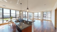 This well-appointed urban residence offers a thoughtfully designed single-floor layout with an abundance of natural light and city views from nearly every room. The open-concept kitchen and living area serves as the heart of the space, showcasing rich warm-toned cabinetry, granite countertops, and a central island with a built-in sink illuminated by stylish pendant lighting. Stainless steel appliances, including a refrigerator, range, and microwave, complete the kitchen. The living area extends generously from the kitchen, featuring gleaming hardwood floors and a wraparound wall of floor-to-ceiling windows that frame panoramic urban views and flood the space with natural light. The bedrooms are carpeted for comfort and each feature large windows with vertical blinds, providing ample natural light and city outlooks. At least one bedroom includes a walk-in closet, adding practical storage. Two full bathrooms mirror each other in design, each outfitted with granite-topped wood vanities, tile flooring, a large mirror, and a shower-tub combination. Throughout the residence, warm wood trim accents the neutral palette, creating a cohesive and inviting atmosphere that balances modern finishes with functional, comfortable living.