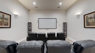 This dedicated home theater room offers a well-appointed cinematic experience within a thoughtfully designed single-level space. The room is anchored by a large wall-mounted projection screen positioned at the front, flanked by tall media storage shelves stocked with an extensive collection of films, making for both a functional and visually striking focal point.

Seating is arranged in two rows of plush, dark leather recliner chairs, each equipped with built-in cup holders for added convenience. The tiered layout ensures clear sightlines from every seat, enhancing the viewing experience for all occupants.

The room's acoustics are supported by a surround sound speaker system, with speakers mounted at strategic points along the walls. Decorative wall sconces provide warm ambient lighting that complements the theater atmosphere, while recessed ceiling lights offer additional illumination when needed. Framed artwork adorns the side walls, adding a refined aesthetic touch.

The interior features a neutral color palette of soft grays and whites, contributing to a clean and immersive environment. Light wood-toned flooring runs throughout, providing a subtle contrast to the dark seating. The space connects to an adjacent hallway through a paneled door, offering easy access while maintaining the room's dedicated, enclosed feel.