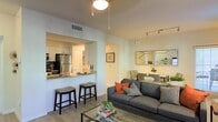 This single-floor residence offers a thoughtfully designed open-concept layout that seamlessly connects the kitchen, dining, and living areas into one cohesive and functional living space. The kitchen is outfitted with stainless steel appliances, including a refrigerator and range, and features granite countertops with a raised breakfast bar accompanied by bar stools — ideal for casual dining or entertaining. White cabinetry and ample counter space contribute to a clean, modern aesthetic. A pass-through design opens the kitchen directly to the rest of the living area, enhancing the sense of flow and connectivity throughout. The dining area, positioned just beyond the kitchen, provides a dedicated space for seated meals and is accented by a decorative mirror that adds depth and brightness to the room. The living room anchors the space with a plush grey sofa dressed in warm accent pillows, a glass coffee table, and a patterned area rug over light wood-style flooring. Track lighting and flush-mount ceiling fixtures provide layered illumination throughout. The neutral color palette, combined with light walls and consistent flooring, creates a bright, welcoming ambiance that feels both practical and refined.