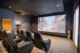 This private movie theater in Memphis, Tennessee, is thoughtfully designed to deliver an immersive cinematic experience within an intimate, upscale setting. The room is enveloped in deep, dark-toned walls and a dark ceiling, creating a focused viewing environment that minimizes light distraction and enhances on-screen contrast. Recessed ceiling lighting provides subtle ambient illumination throughout the space without compromising the theater atmosphere.

The centerpiece of the room is a large projection screen mounted on the front wall, capable of displaying a full cinematic image with vivid clarity. Seating is arranged in tiered rows of black leather recliner-style chairs equipped with integrated cup holders, offering both comfort and optimal sightlines to the screen from every position. The elevated rear row ensures unobstructed views for all guests.

A textured, striped carpet covers the floor, adding warmth and sound-dampening qualities to the room. Along the rear wall, two louvered wooden doors provide access, while a stylish herringbone-patterned credenza topped with a black table lamp adds a decorative touch. A frosted-glass entry door is also present near the screen wall. The overall design balances functionality with refined aesthetics, making this theater well-suited for private screenings and entertaining.