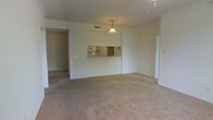 This single-floor residence offers a practical and functional layout centered around an open living area. The spacious carpeted living room serves as the heart of the home, finished in neutral tones that lend a clean, understated look throughout. A defining architectural feature is the pass-through window connecting the living area to the kitchen, enhancing the sense of openness and allowing for easy interaction between the two spaces. A classic pendant light hangs above the pass-through, providing both focused illumination and a subtle decorative touch. The kitchen, visible through the opening, is equipped with wood-toned cabinetry and a built-in microwave, reflecting a straightforward and efficient design. Ceiling-mounted HVAC vents are integrated neatly into the layout, ensuring year-round comfort. An entryway with tile flooring is visible to the side, providing a practical transition zone at the home's entrance. A hallway leads further into the residence, suggesting additional private rooms beyond. The overall palette of soft whites and warm beige tones creates a calm, neutral ambiance throughout, making the space well-suited for a variety of furnishing styles and personal tastes.