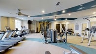 Located at 1015 Nutt Street in Wilmington, NC, this fitness center offers a thoughtfully designed workout environment that combines functionality with modern aesthetics. The space features a distinctive teal and neutral color scheme carried across the flooring and ceiling accents, creating a cohesive and energizing atmosphere throughout. Recessed lighting and ceiling fans are evenly distributed across the ceiling, ensuring a well-lit and comfortable environment for all types of exercise. The main workout floor is generously proportioned, accommodating a comprehensive collection of Matrix-brand strength training machines arranged in an open, accessible layout. A dedicated free weights section lines one wall, complete with a full dumbbell rack and wall-to-wall mirrors that enhance the sense of space and allow for proper form checks. Cardio equipment, including treadmills and elliptical machines, is positioned along the perimeter, offering ample room for users to move freely between stations. An exposed brick accent wall adds a warm, industrial character to the space, balancing the sleek modern equipment. Built-in shelving provides additional storage, and the overall layout is spacious enough to comfortably serve multiple users simultaneously. This is a well-equipped, professionally appointed fitness facility suited for a full range of workout routines.