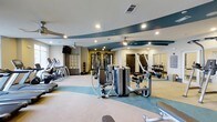 Located at 1015 Nutt Street in Wilmington, NC, this fitness center offers a thoughtfully designed workout environment that combines functionality with modern aesthetics. The space features a distinctive teal and neutral color scheme carried across the flooring and ceiling accents, creating a cohesive and energizing atmosphere throughout. Recessed lighting and ceiling fans are evenly distributed across the ceiling, ensuring a well-lit and comfortable environment for all types of exercise. The main workout floor is generously proportioned, accommodating a comprehensive collection of Matrix-brand strength training machines arranged in an open, accessible layout. A dedicated free weights section lines one wall, complete with a full dumbbell rack and wall-to-wall mirrors that enhance the sense of space and allow for proper form checks. Cardio equipment, including treadmills and elliptical machines, is positioned along the perimeter, offering ample room for users to move freely between stations. An exposed brick accent wall adds a warm, industrial character to the space, balancing the sleek modern equipment. Built-in shelving provides additional storage, and the overall layout is spacious enough to comfortably serve multiple users simultaneously. This is a well-equipped, professionally appointed fitness facility suited for a full range of workout routines.