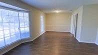 This single-floor space offers a clean, functional layout characterized by a warm and inviting aesthetic. The main living area showcases rich, dark hardwood flooring that runs throughout, providing a striking contrast against the soft, neutral-toned walls. A classic chair rail detail runs along the perimeter of the room, adding a subtle architectural touch that lends a sense of traditional character to the space.

Natural light filters in generously through large windows fitted with white horizontal blinds, brightening the interior and creating an airy atmosphere. The textured ceiling adds depth overhead, while flush-mount ceiling lighting ensures the space remains well-lit at any time of day.

White trim and baseboards frame the room cleanly, complementing the overall light color palette and contributing to a crisp, well-maintained appearance. A set of bifold doors, likely leading to a closet or utility space, is visible along one wall, offering practical storage solutions without compromising the open feel of the room.

The layout is open and versatile, lending itself well to a variety of uses and furniture arrangements. The combination of durable flooring, ample natural light, and thoughtful architectural detailing makes this a comfortable and adaptable single-level space.